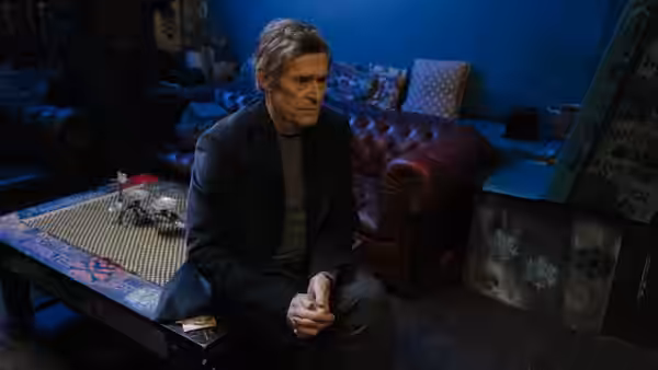 Ed Saxberger (Willem Dafoe) seated on the floor in a blue room
