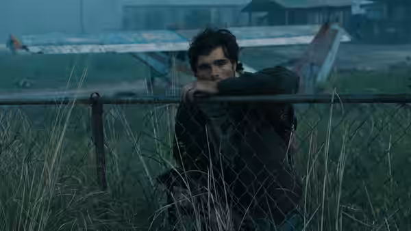 Hig (Jacob Elordi) leaning on a chain-link fence in front of an airplane