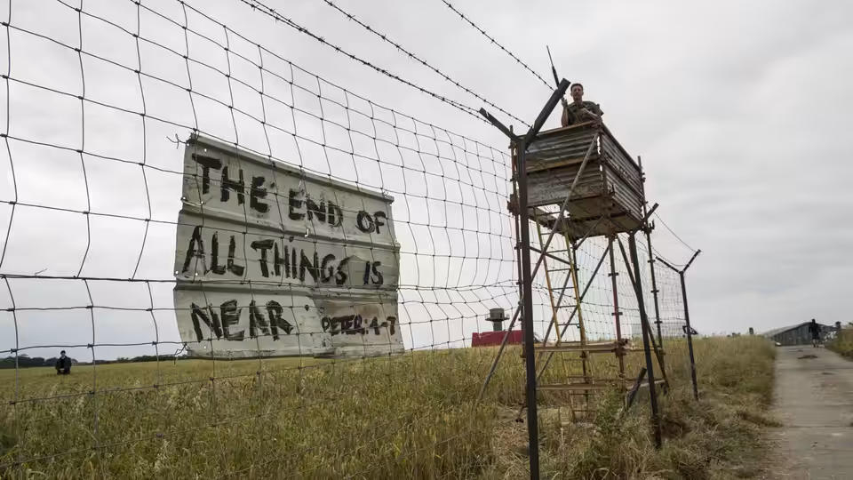 A guard watches from a wooden tower behind a barbed-wire fence bearing a warning sign