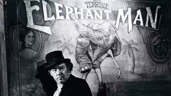 A man in a top hat stands before a large Elephant Man poster
