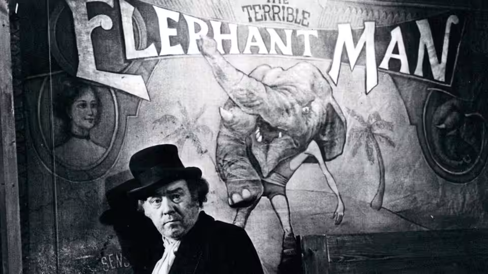 A man in a top hat stands before a large Elephant Man poster