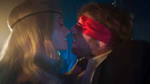 A blonde woman leans close to a man wearing a red blindfold