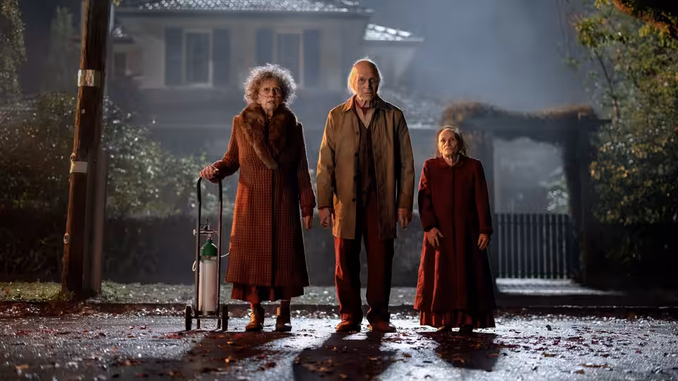 Three elderly people stand together on a wet path outside a house at night