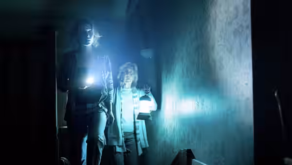 Two women walk through a dark hallway holding flashlights and looking ahead