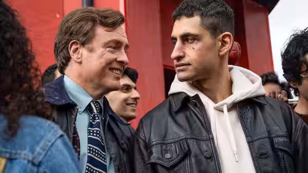 Naz (Amir El-Masry) stands with Frank Warren (Toby Stephens) near a red wall