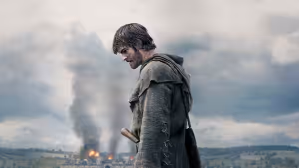 man in tattered cloak stands before a burning settlement under clouds