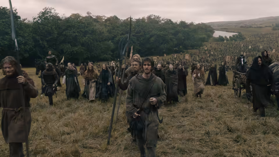 large group of peasants walks across a grassy field carrying weapons