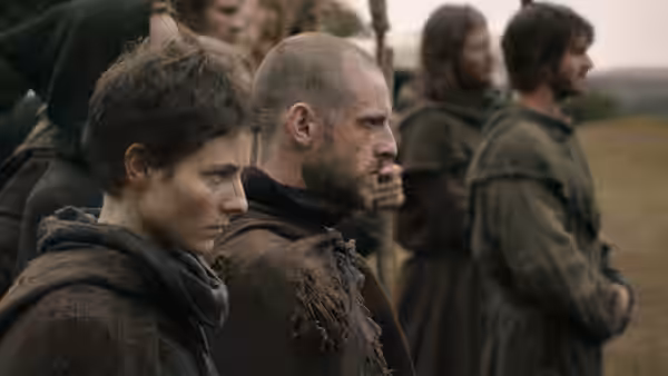 young woman and bald man in rough tunics stand in a field with others behind