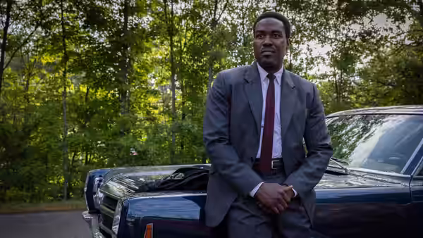 Wayne Strider (Yahya Abdul-Mateen II) leans against a blue car in woods
