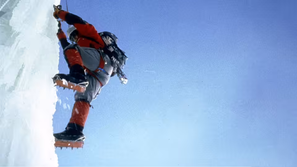 Climber in red jacket scaling steep ice wall with crampons and axe