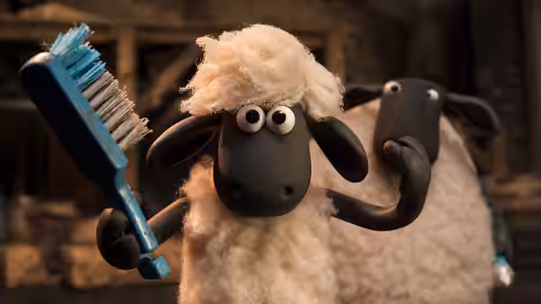 Shaun holds a blue brush while another sheep stands behind him indoors