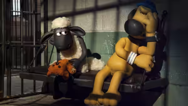 Shaun sits holding a stuffed toy beside Bitzer on a bench in a barred room
