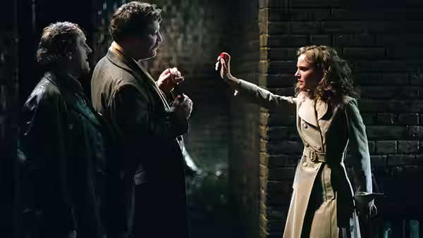 Woman in trench coat holds up red rose before two men in dim brick corridor