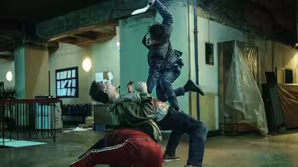 A woman kicks a man held by another in an empty warehouse