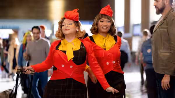 Two flight attendants in red and yellow pull luggage through a busy terminal