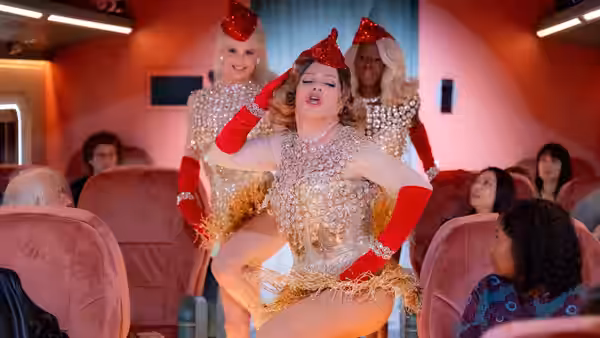 A performer in gold sequins dances inside an airplane cabin with others behind
