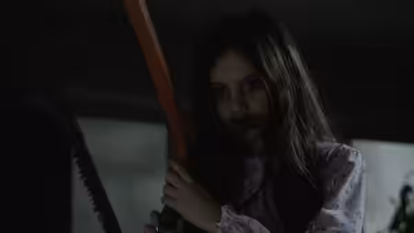 A girl grips a large chainsaw and stares ahead in shadow