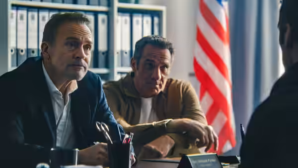 Two men seated at a desk in an office with an American flag behind them