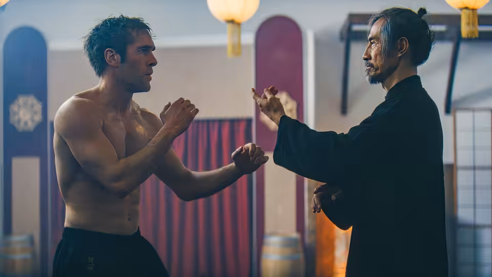Shirtless man and older man in black robe facing off in fighting stances