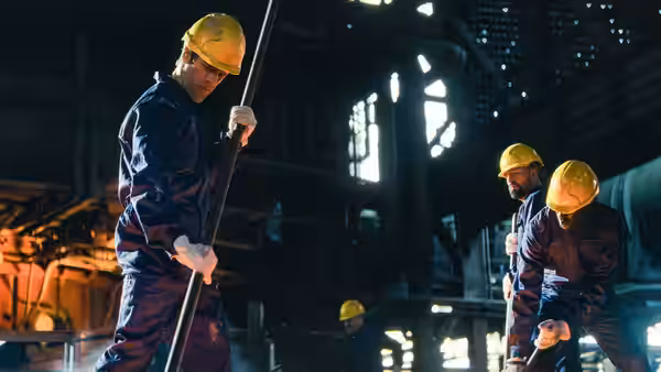 Three men in hard hats face off with long poles inside an industrial facility