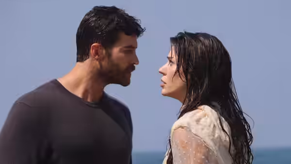 A dark-haired man and a woman face each other beside the sea