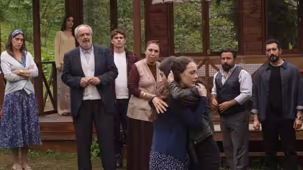 A woman embraces a younger person outside a wooden building as relatives watch