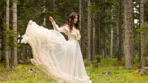 A woman in a flowing white dress lifts its train among the trees