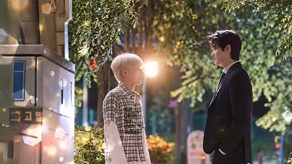 A white-haired young man faces a dark-haired man in a suit beside a sunlit street