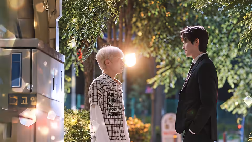 A white-haired young man faces a dark-haired man in a suit beside a sunlit street