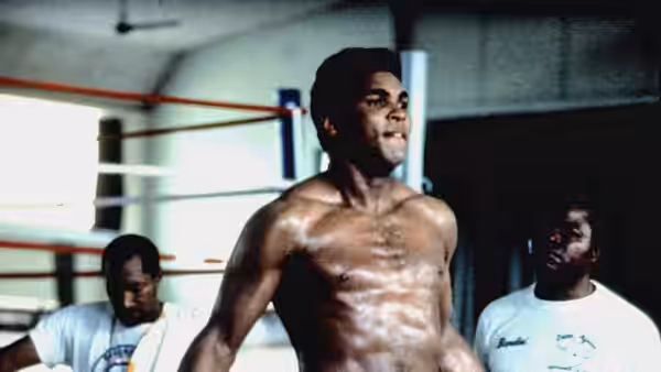 Muhammad Ali standing shirtless inside a brightly lit boxing gym