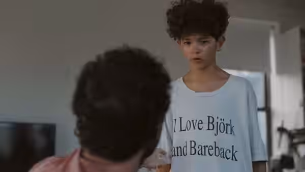 A curly-haired boy stands in a white T-shirt as a seated man looks up at him