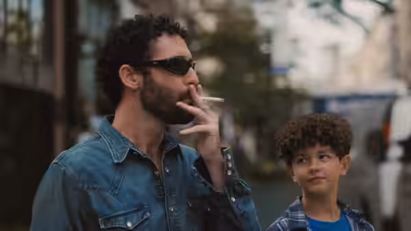 A bearded man smokes on a city sidewalk beside a curly-haired boy