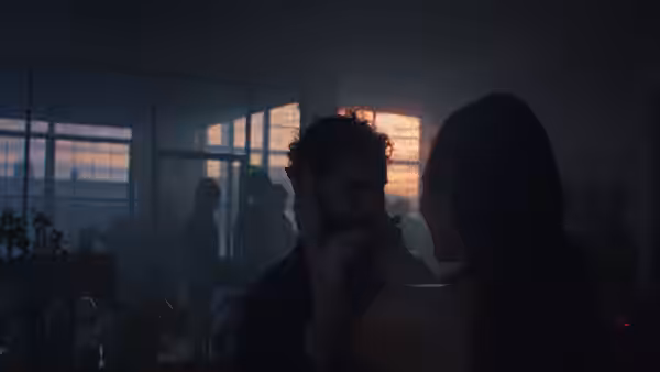 Two figures face each other in a dim apartment as sunset glows through the windows