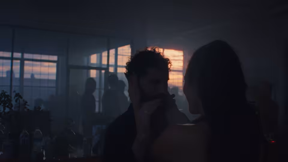 Two figures face each other in a dim apartment as sunset glows through the windows