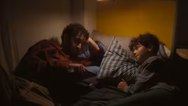 Peter (Jordan Firstman) sits beside a boy on a bed in a dim room