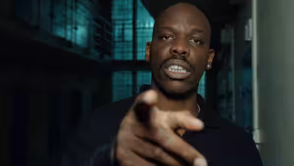 A man in a dark polo shirt points forward in a blue-lit prison corridor