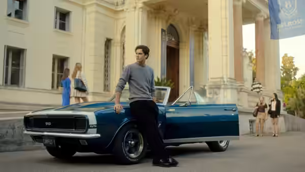 A young man leans against a blue convertible with its door open outside a university building