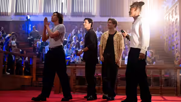 Four young men stand before a seated crowd inside a hall