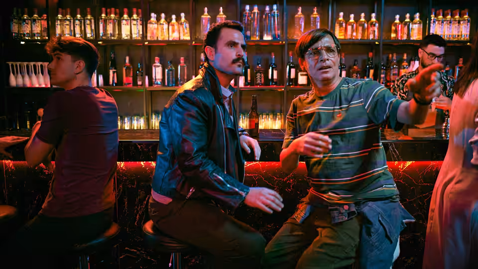 Men seated at a bar counter with bottles behind them one gesturing