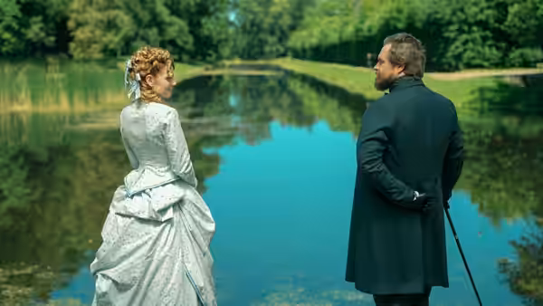 A woman in a pale gown and a man with a cane stand beside a pond