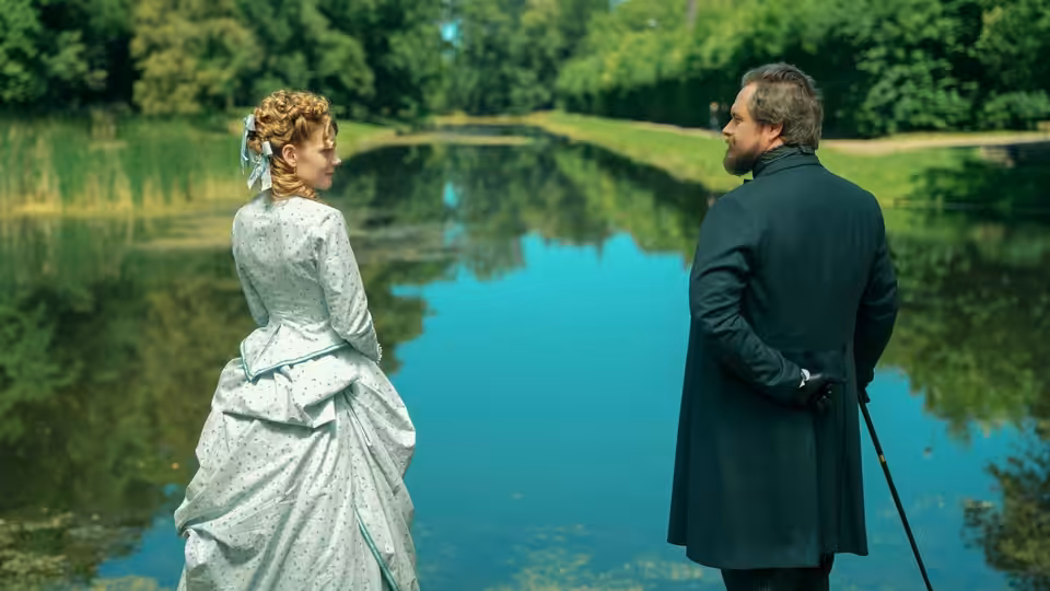 A woman in a pale gown and a man with a cane stand beside a pond