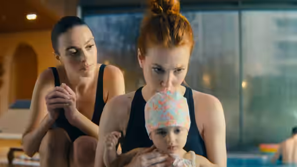 Two women in black swimsuits by an indoor pool, one holding a baby