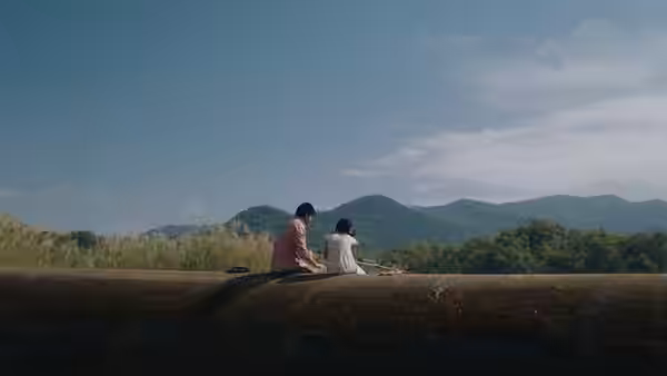 Two people sit on a low embankment facing a range of green mountains