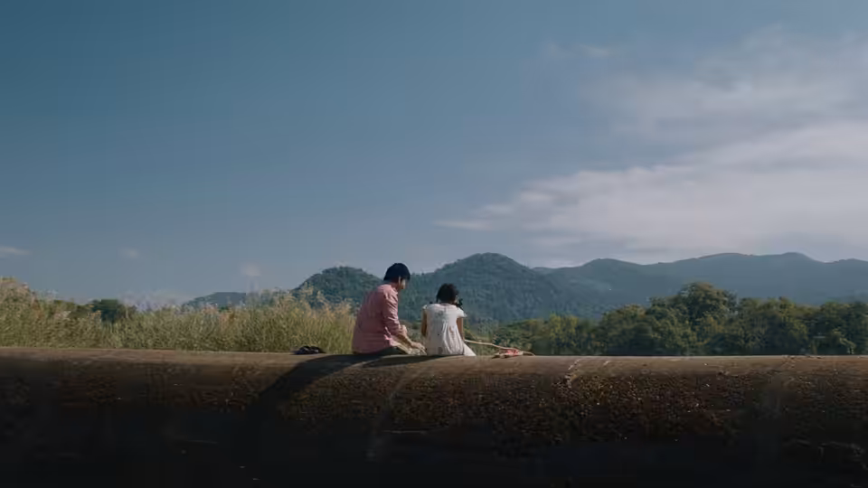 Two people sit on a low embankment facing a range of green mountains