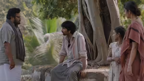 A seated man talks with another man and two women beneath a large tree