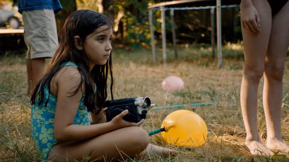 A young girl in a swimsuit holding a video camera on grass