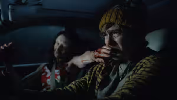 Man in yellow beanie eats from bloody hand in car with injured woman behind