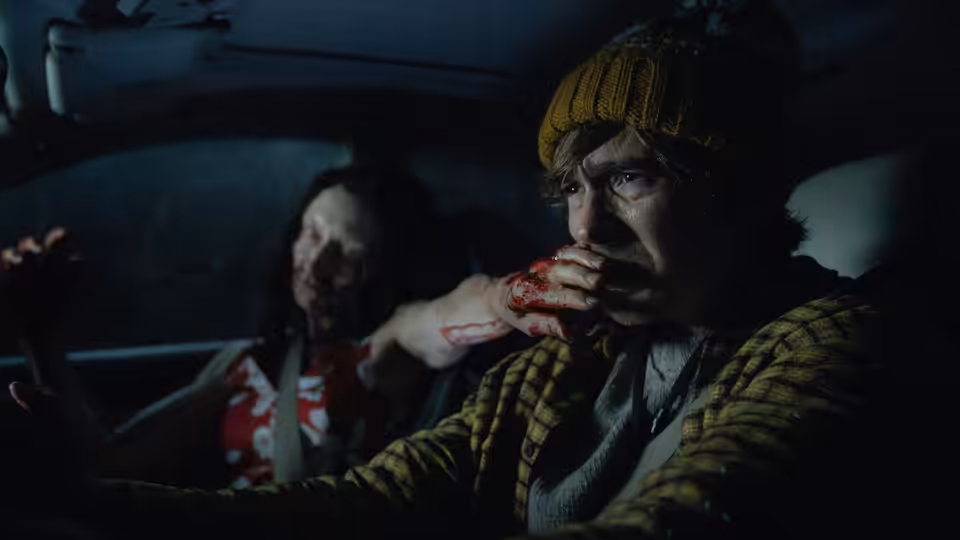 Man in yellow beanie eats from bloody hand in car with injured woman behind