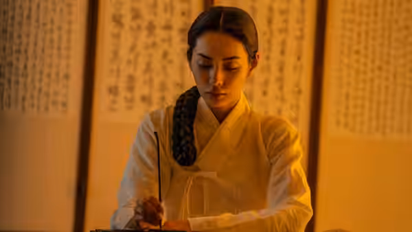 A braided woman writes at a desk before hanging calligraphy scrolls