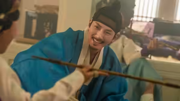 A smiling man in blue robes leans toward a seated figure holding a wooden rod